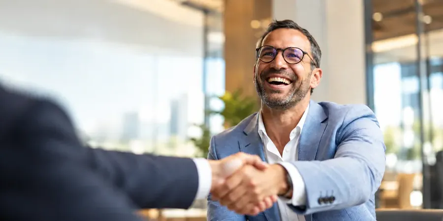 A smiling man in a blazer shaking hands across a meeting table