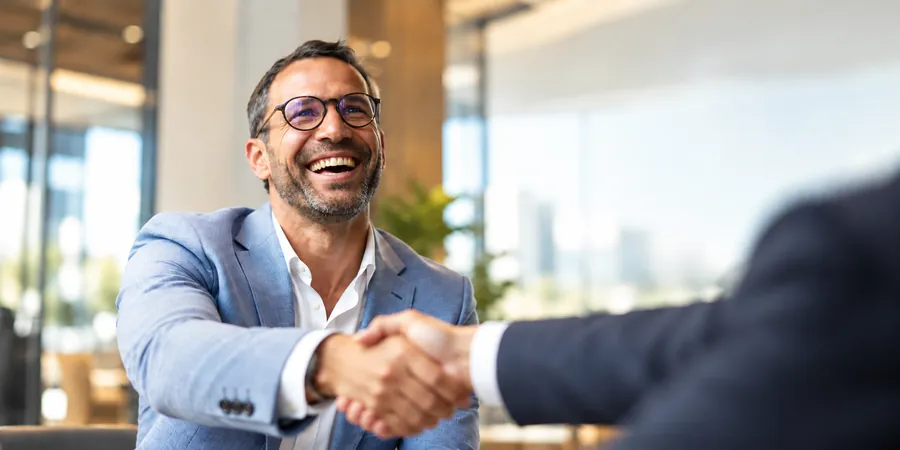 A smiling man in a blazer shaking hands across a meeting table