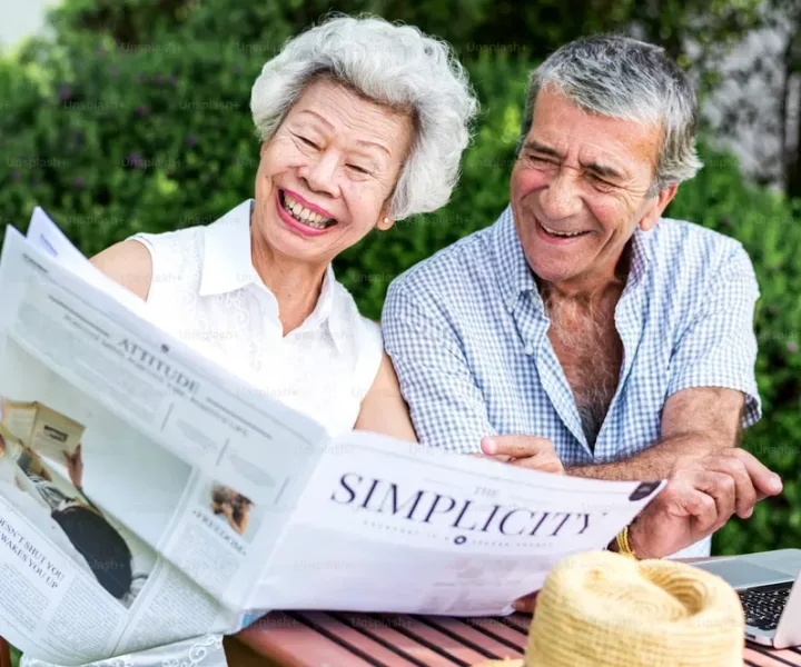 An older couple sitting together outdoors, reading and laughing