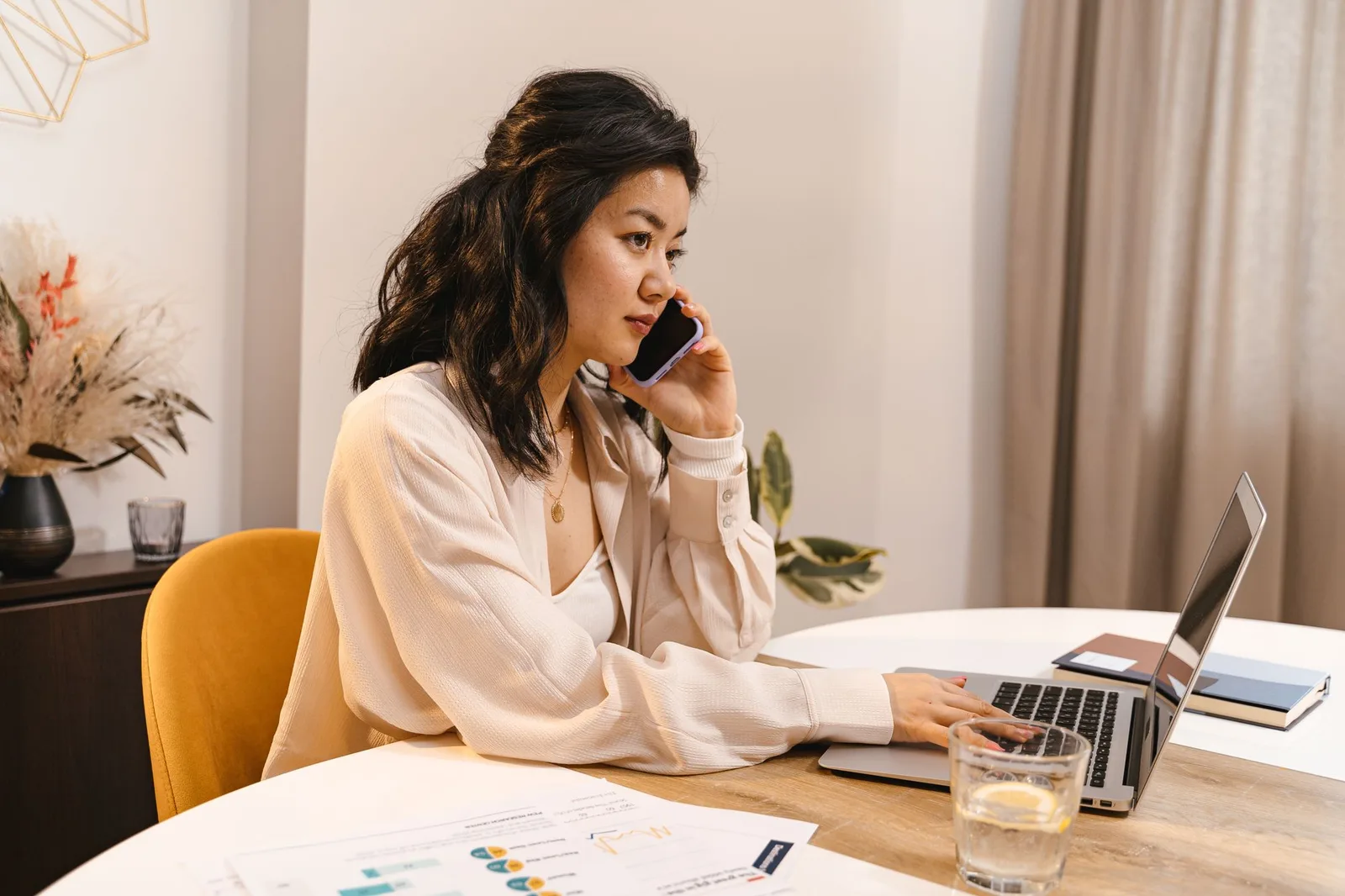 Woman on a phone call at a home desk with a laptop and notes