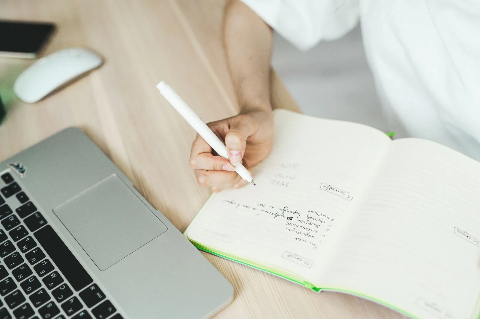 Hand writing a list in a notebook beside a laptop